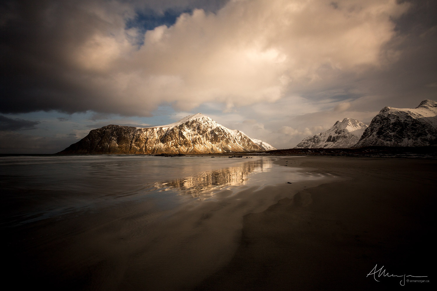 Snow capped mountains reflected in a calm, wet sand beach at sunset after a winter storm.