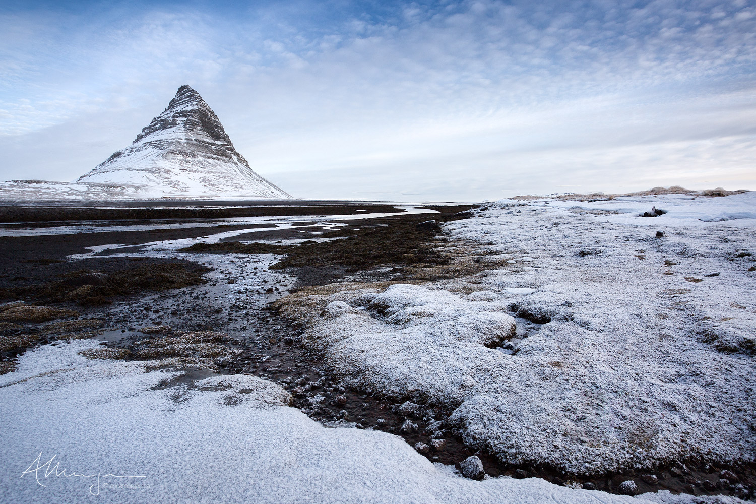 The iconic Kirkjufell mountain in Iceland taken from the nearby black sand beach at low tide with a fresh snowfall gently blanketing the seaweed.