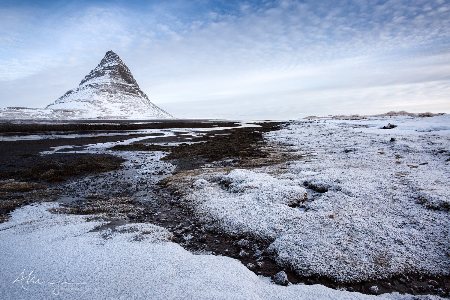 Powdered Sugar The iconic Kirkjufell mountain in Iceland taken from the nearby black sand beach at low tide with a fresh snowfall gently blanketing the seaweed.